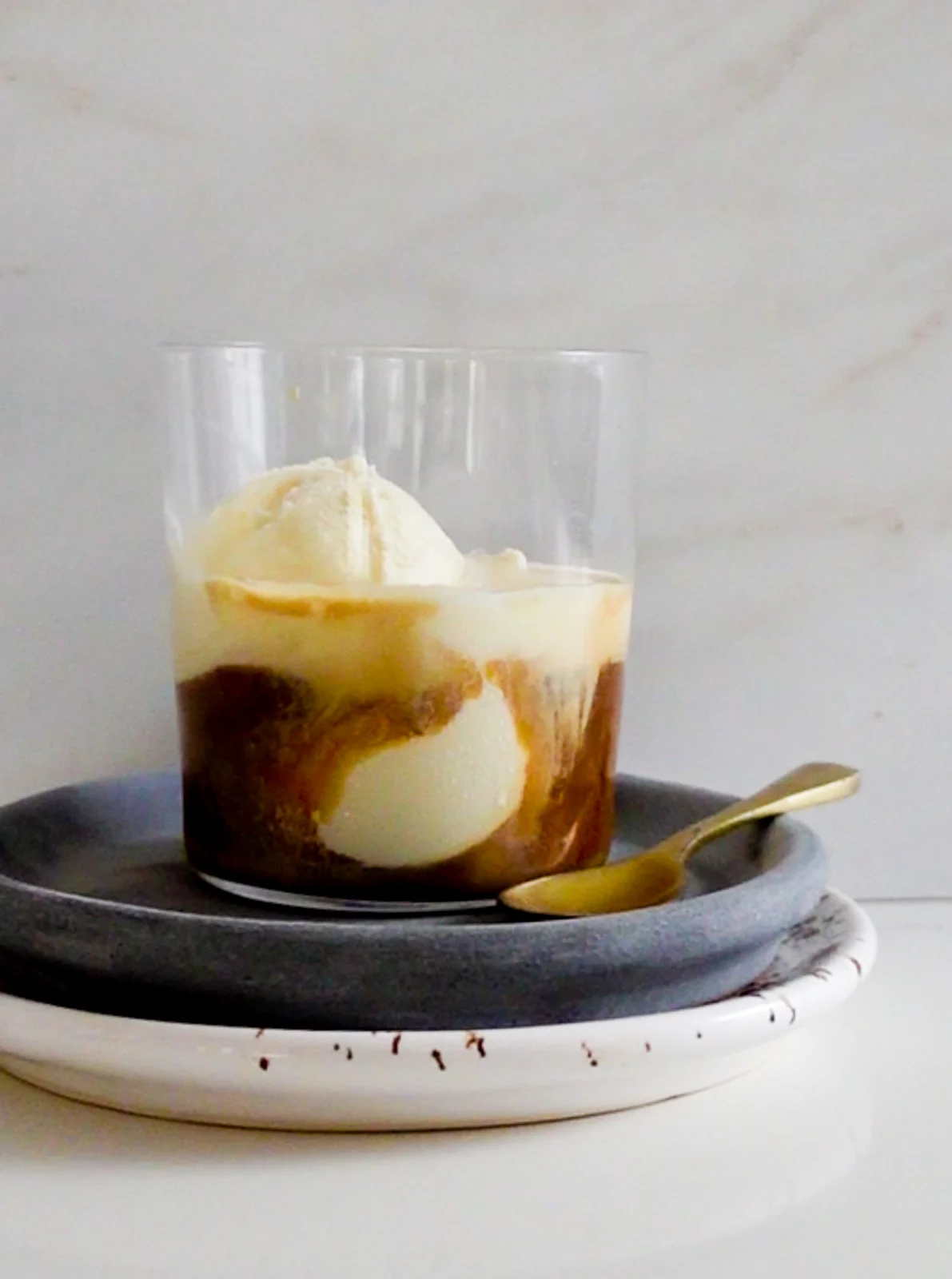 A glass cup filled with creamy vanilla ice cream, elegantly transformed into an affogato with a hot espresso pour, creates a swirling effect on a gray plate atop a white one. A small gold spoon rests nearby. The light marble surface background adds to the dessert's minimalist elegance.